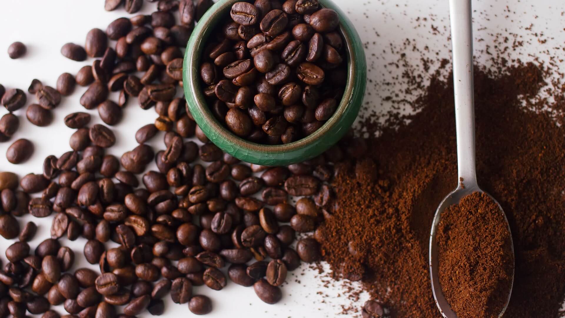 Coffee beans in a cup and spilling out onto a table next to coffee grounds on the table and in a metal spoon.