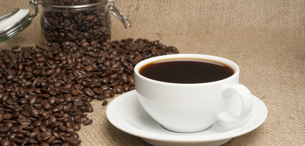 A cup of coffee in a white cup on a table that also has coffee beans laid out on the table and in a glass jar