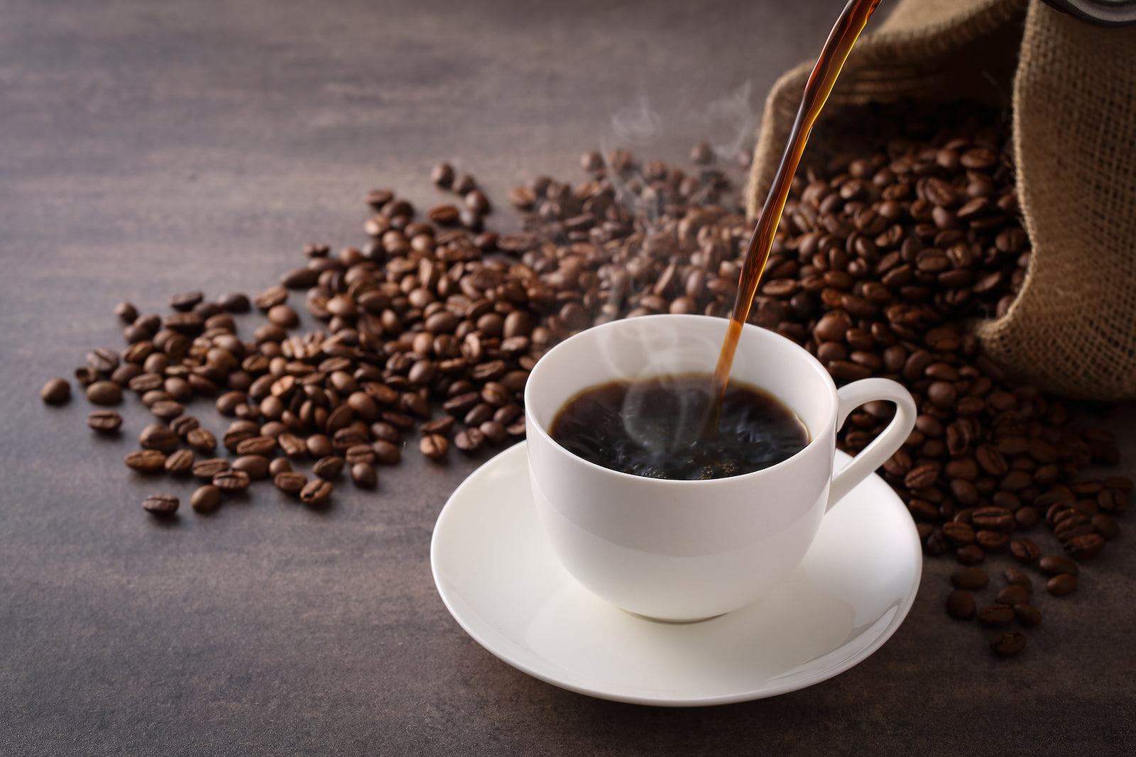A white cup is being filled with steaming coffee on a saucer, surrounded by scattered coffee beans and a burlap sack on a dark surface.