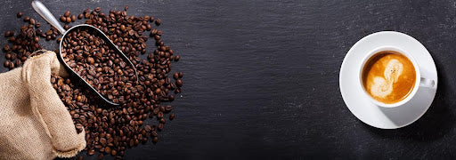 Unroasted coffee beans spill from a metal scoop next to a burlap sack. A white cup holds frothy espresso, resting on a dark, textured surface.