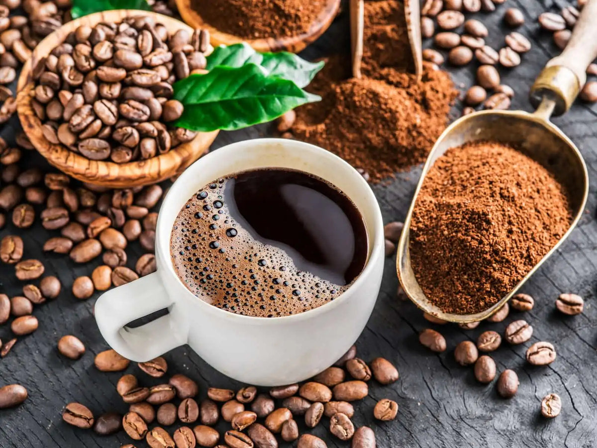 A table with coffee beans, ground coffee, green leaves, and a white mug with freshly brewed coffee