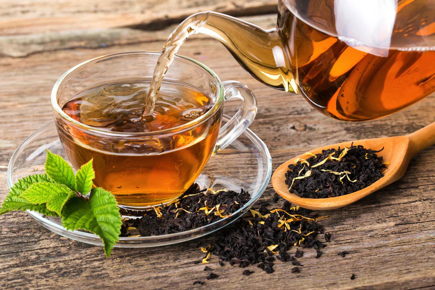 Tea in a clear teapot being poured into a clear mug sitting on a saucer on top of black loose leaf tea