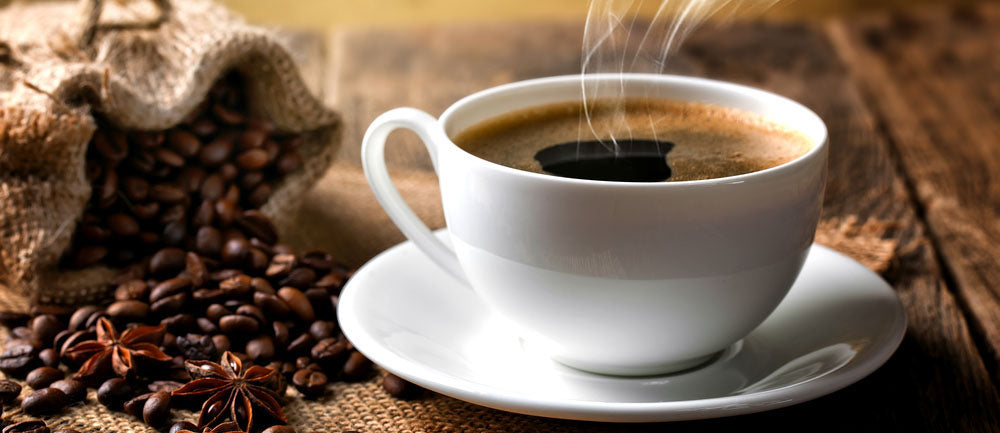 A steaming cup of freshly brewed coffee in a white mug placed next to a bag full of spilled over coffee beans