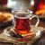 A steaming glass of tea sits on a clear saucer with cloves and cookies, placed on a brown textured cloth. In the background, a warm, blurred environment is visible.