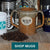 Double handled tan mug with Mystic Monk Coffee logo sitting on wooden board with coffee beans in foreground and assorted mugs partially visible in the background. Text in image reads Shop Mugs